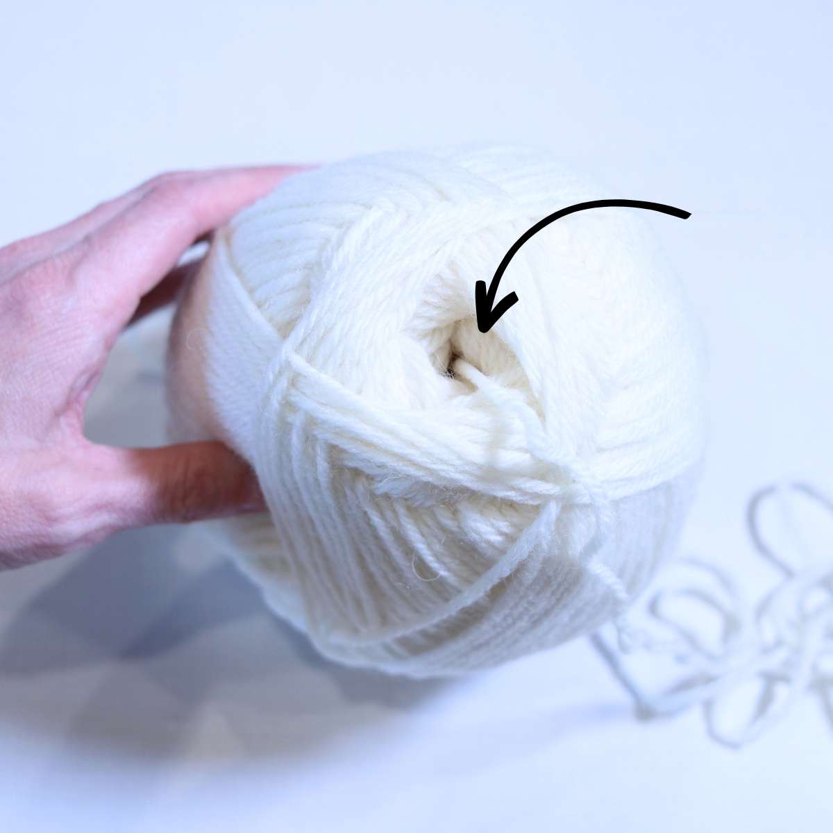 A womans hand is holding a skein of cream colored yarn towards the camera to demonstrate where to pull the yarn from when making DIY wool dryer balls. A black arrow is pointing to the center of the yarn ball.