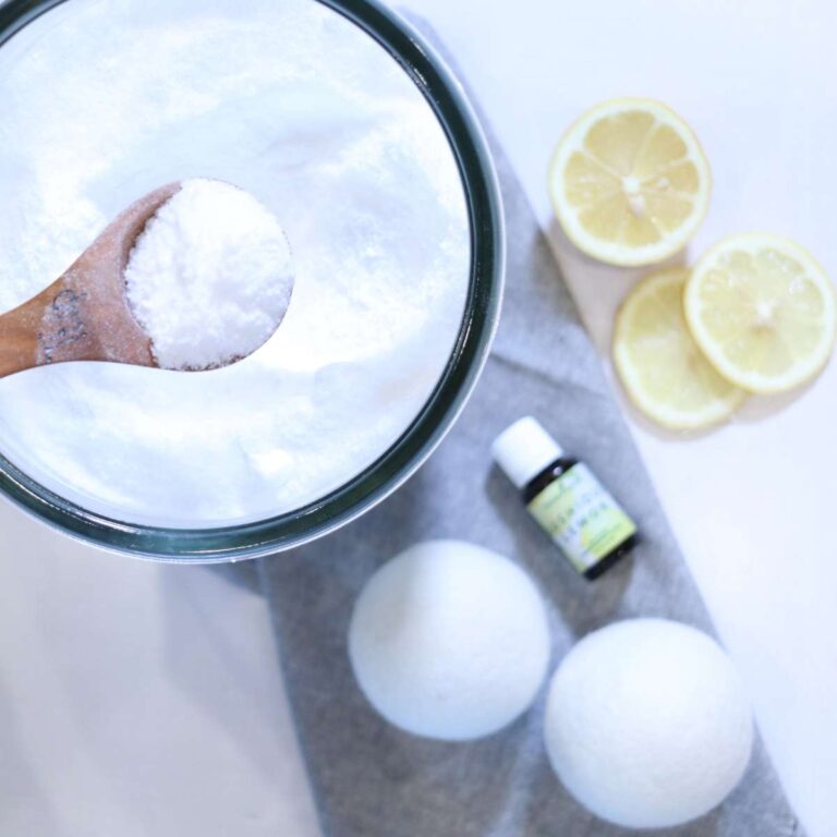 A wooden measuring spoon filled with DIY laundry scent booster. Two white wool felted dryer balls, a bottle of lemon essential oil and lemon slices rest neatly near the clear glass jar.