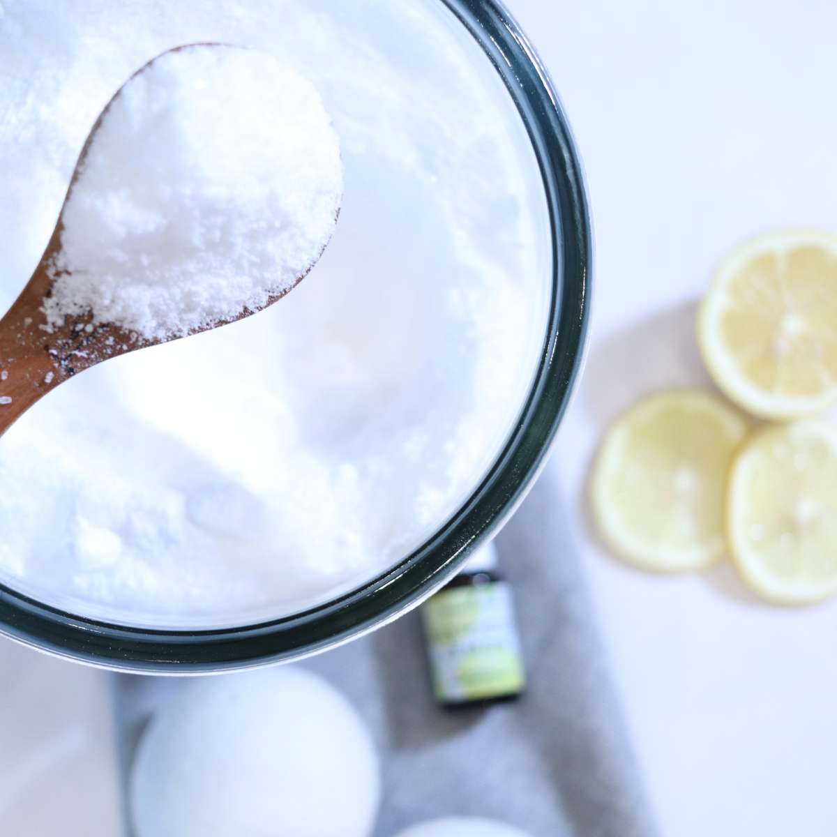 A wooden spoon has scooped some DIY laundry scent booster recipe that is ready to be added to a wash load. Two felted wool dryer balls, a bottle of lemon essential oil and lemon slices are sitting next to the clear glass storage jar.