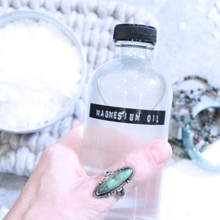 A Womans hand wearing a turquoise ring is holding on to a clear glass bottle full of homemade magnesium oil. A small bowl of magnesium flakes and a stack of turquoise bracelets sits on the table in the background.