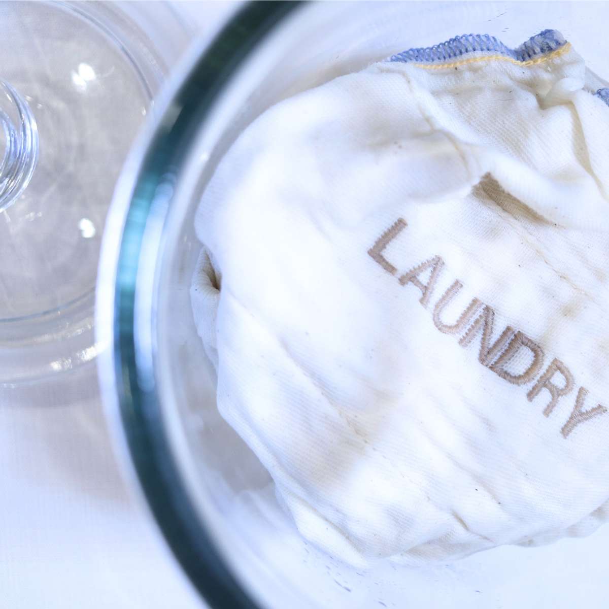 A clear glass jar full of ready to use homemade dryer sheets. The dryer sheets are a crisp white with blue stitching along the edges. The word laundry has been embroidered on the front of the cloth fabric sheets.