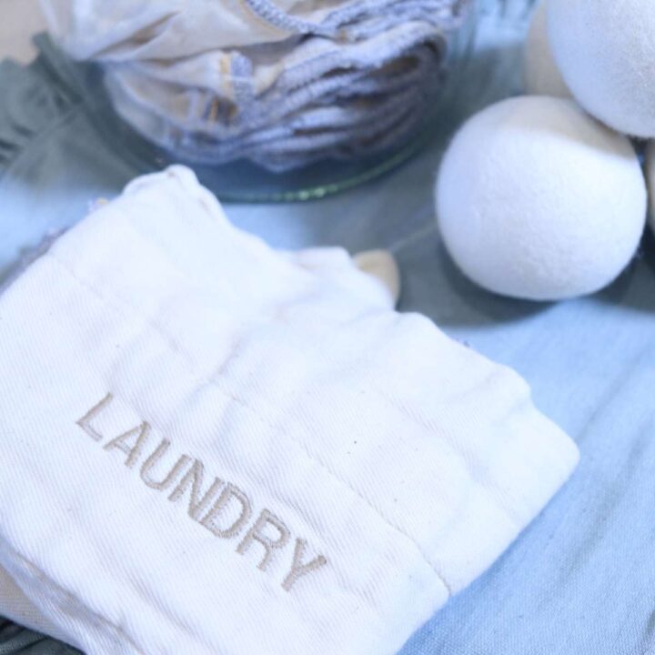 Some DIY Non-Toxic Dryer Sheets folded in a neat stack ready for use. The homemade dryer sheets are sitting next to a clear jar that also has dryer sheets inside. Some white wool dryer balls are resting on the table nearby.