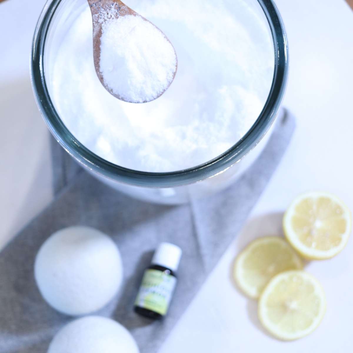 A clear glass storage jar full of white powdered DIY laundry scent booster. A wooden spoon has scooped some of the mixture. Two white wool felted dryer balls, sliced lemon and a bottle of lemon essential oil are resting on the table near the jar.