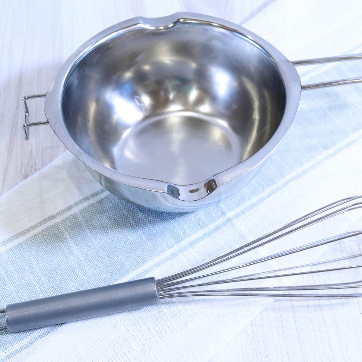 A small silver pot with 2 pour spouts is a store-bought double boiler. A silver metal whisk sits next to the pot.