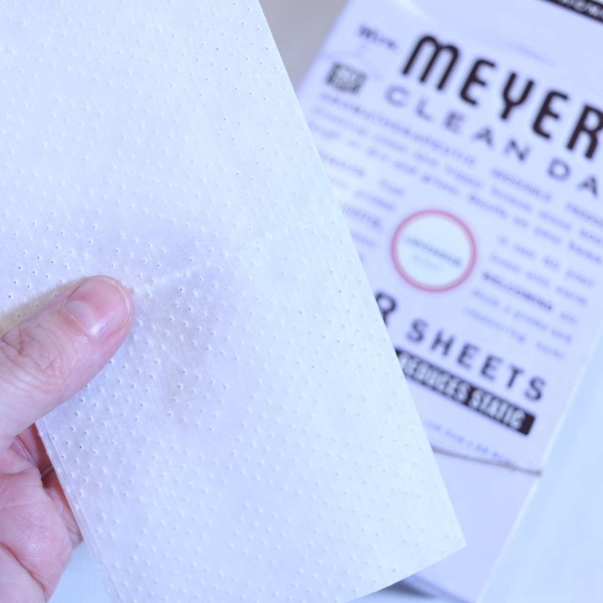 A womans hand is holding onto a white folded store bought dryer sheet to show texture. The lavender box for the dryer sheets rests in the background.