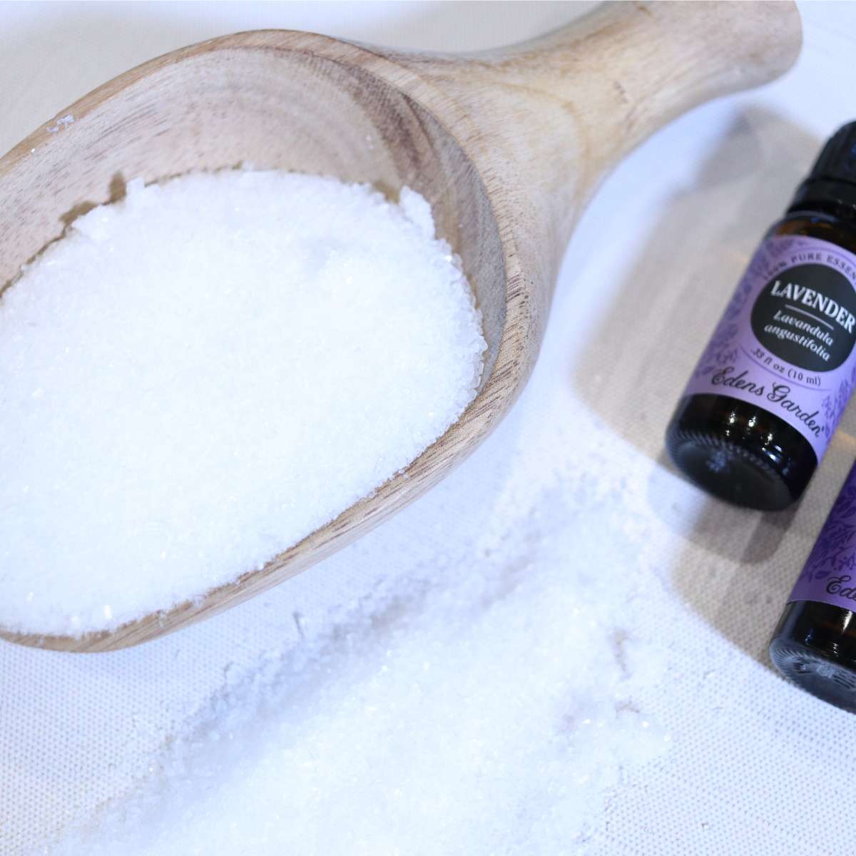 A wooden scoop filled with epsom salt for a DIY epsom salt based DIY laundry scent booster recipe. Two bottle of lavender essential oils are near a small pile of spilled epsom salt on the table top.