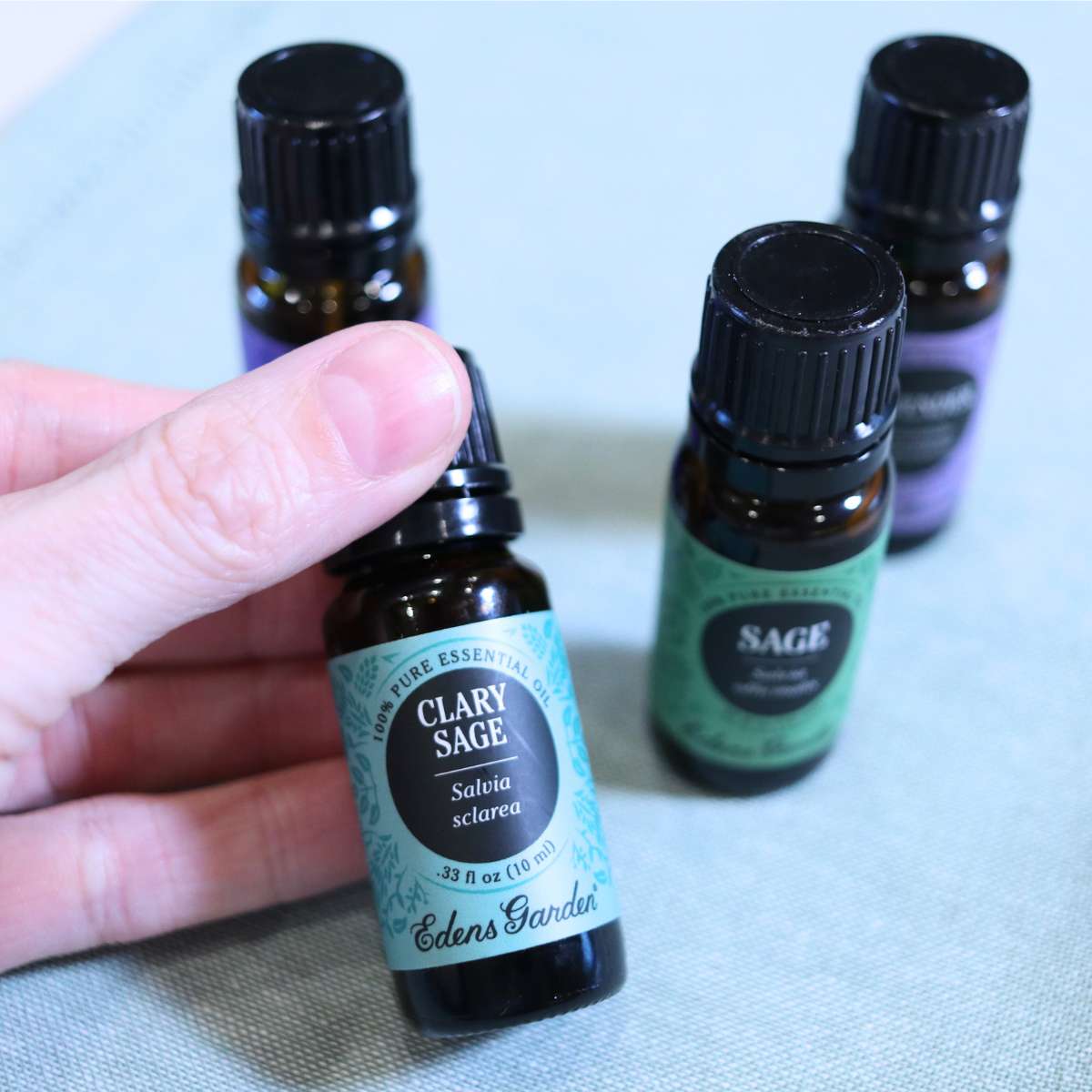 A hand is holding a bottle of clary sage essential oil while three other bottles of different scented oils sits nearby. These essential oils are being used for DIY cleaning recipes.