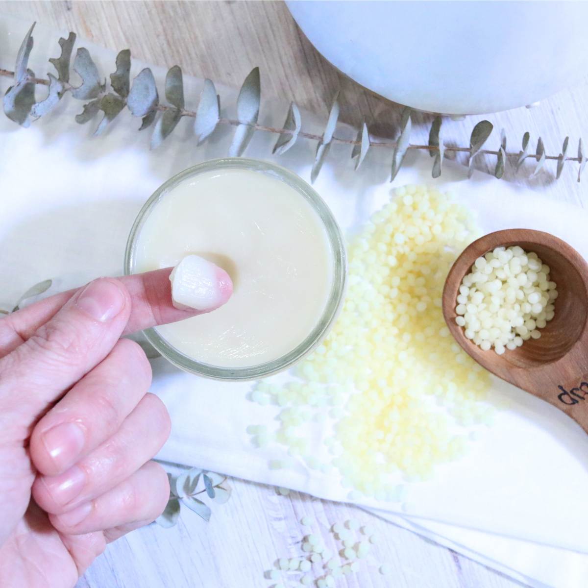 A womans index finger has a small amount of homemade vapor rub on the tip to demonstrate consistency of her all-natural vapor rub she just made. A wooden measuring spoon of beeswax pellets and a sprig of eucalyptus can be seen in the background of the photograph. 