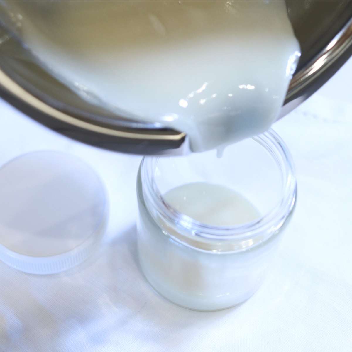 A silver double boiler with pour spout is carefully pouring homemade deodorant without baking soda into a clear glass jar. A white plastic lid for the jar sitting nearby.