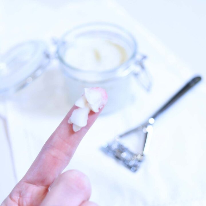 A womans index finger has a generous pea sized amount of homemade natural deodorant without baking soda on it. A silver zero waste razor and small clear glass jar filled with homemade deodorant are in the background.