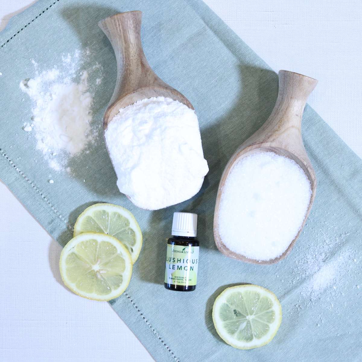 Two wooden scoops filled with epsom salt and baking soda powder for homemade laundry scent booster recipe. A bottle of lemon essential oil and lemon slices are near the wooden scoops.