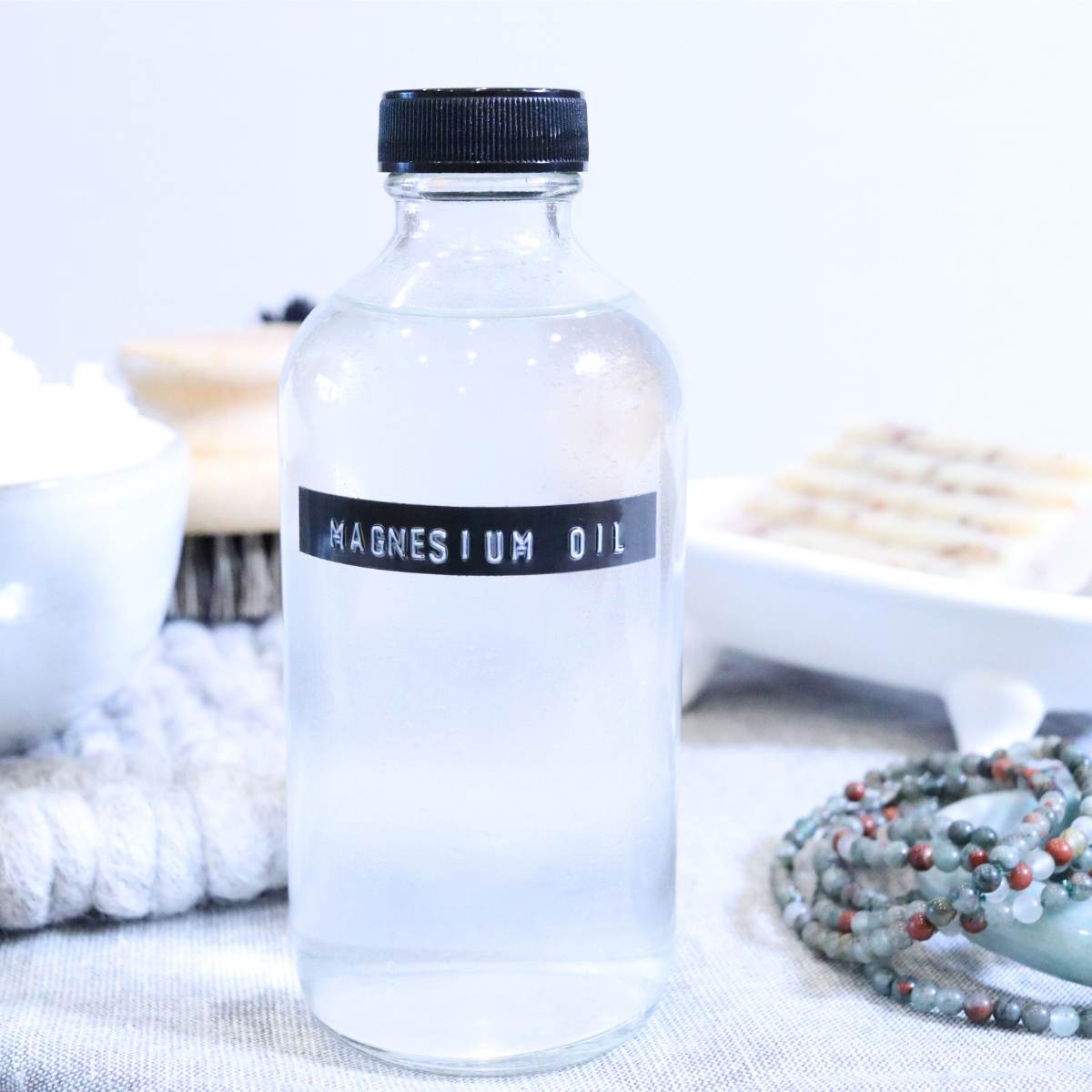 A fresh bottle of magnesium oil that will be gifted to a friend expecting her first baby. A homemade bar soap and a dry brush are seen in the backdrop next to a small bowl of white magnesium flakes. The clear bottle of magnesium oil has a small embossed label on the front.