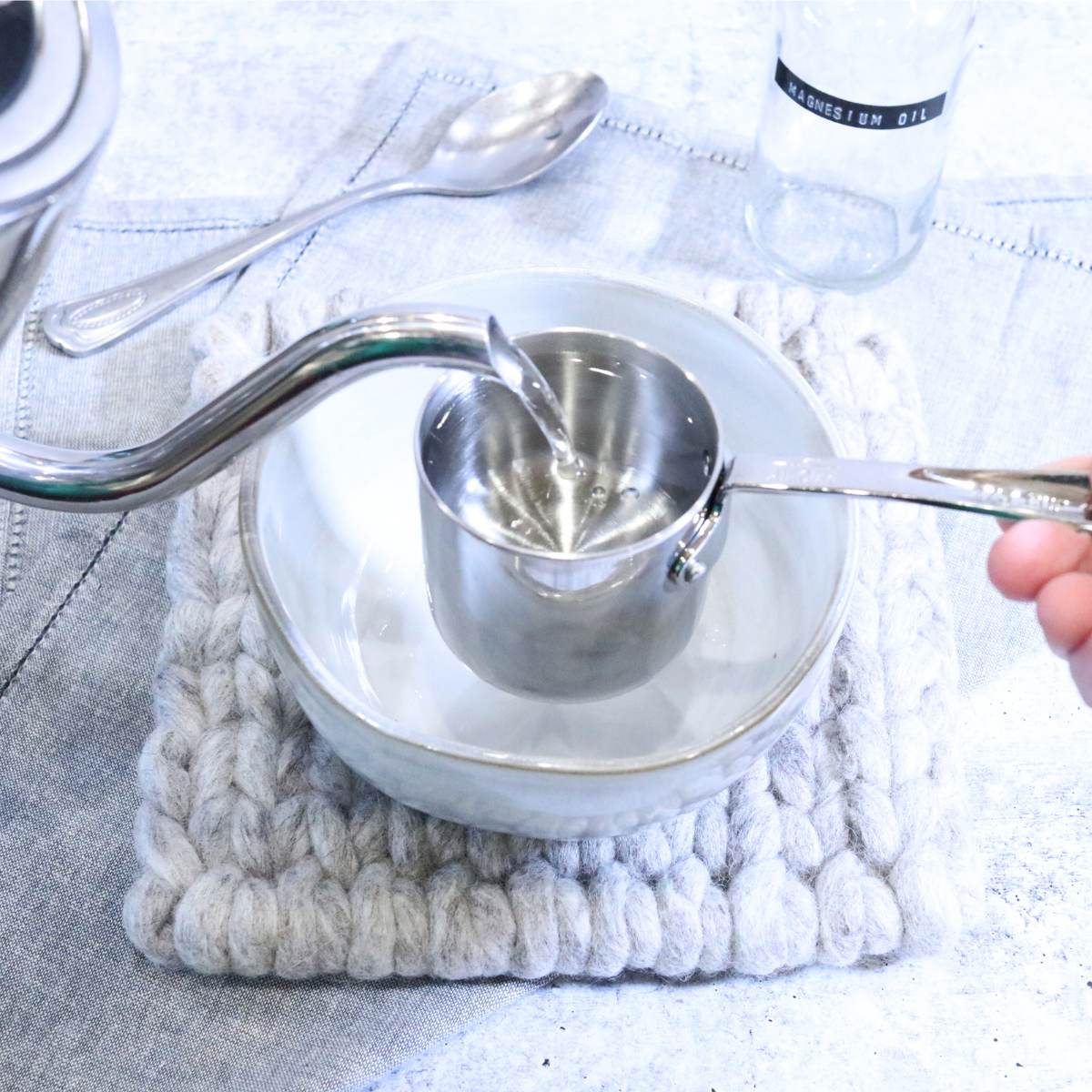 A small silver tea kettle spout pouring water into a silver measuring cup for the DIY magnesium oil recipe. A silver spoon and empty glass storage jar are in the background of the photograph.