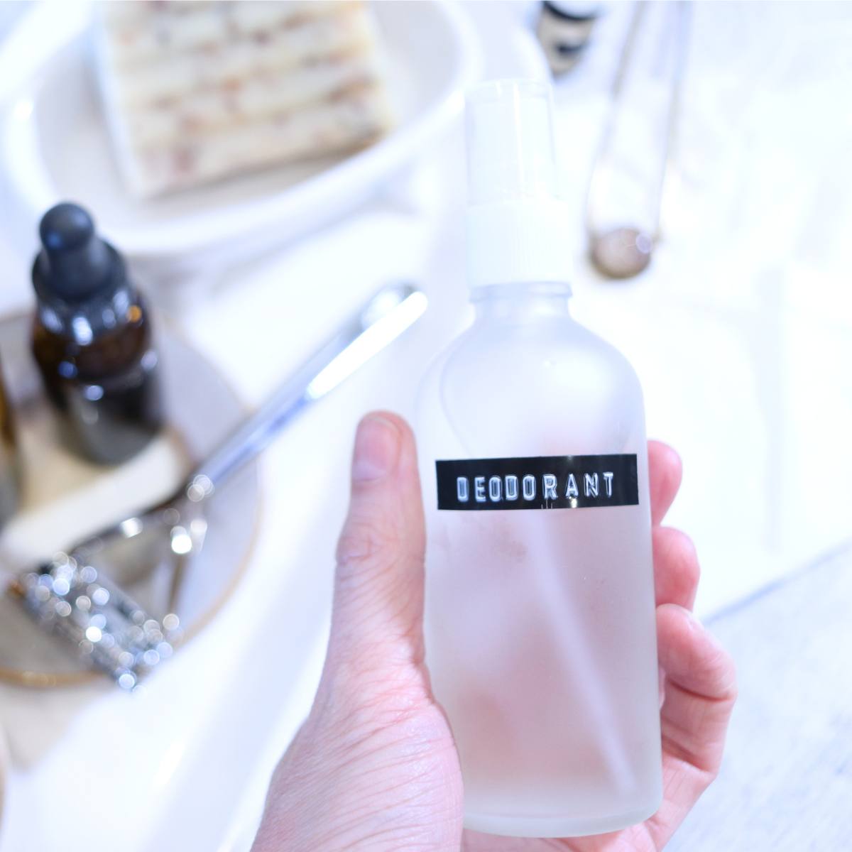 A womans hand is holding a clear glass spray bottle full of homemade magnesium deodorant. There are amber glass bottles of carrier oils, a zero waste silver razor and a bar of patchouli soap in the background of the photograph.