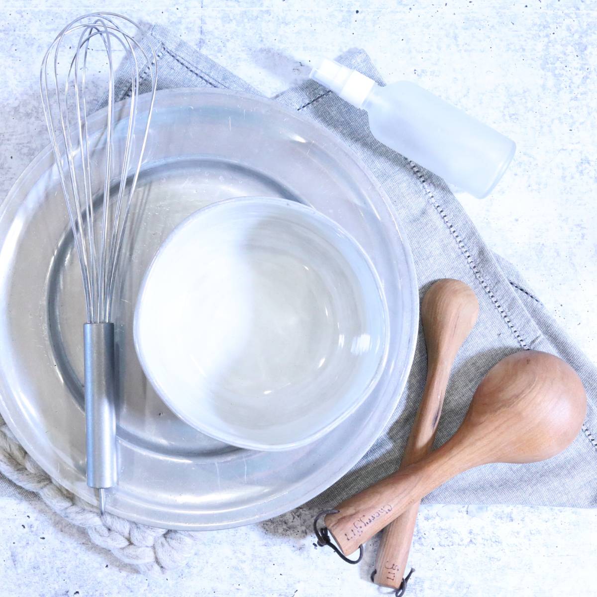 Supplies used for making homemade magnesium deodorant. A silver platter has a medium bowl used for mixing ingredients. A silver metal whisk, wooden measuring cup and measuring spoon and a clear glass spray bottle all resting onto of a gray folded linen cloth resting on a table.