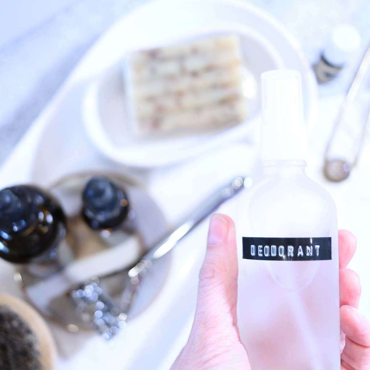 A Womans hand is hold a clear glass spray bottle filled with homemade magnesium deodorant she made. Behind her hand is other bathroom items including a silver zero waste razor, two amber bottles full of oils, a bar of soap, a hair pin and a dry brush.