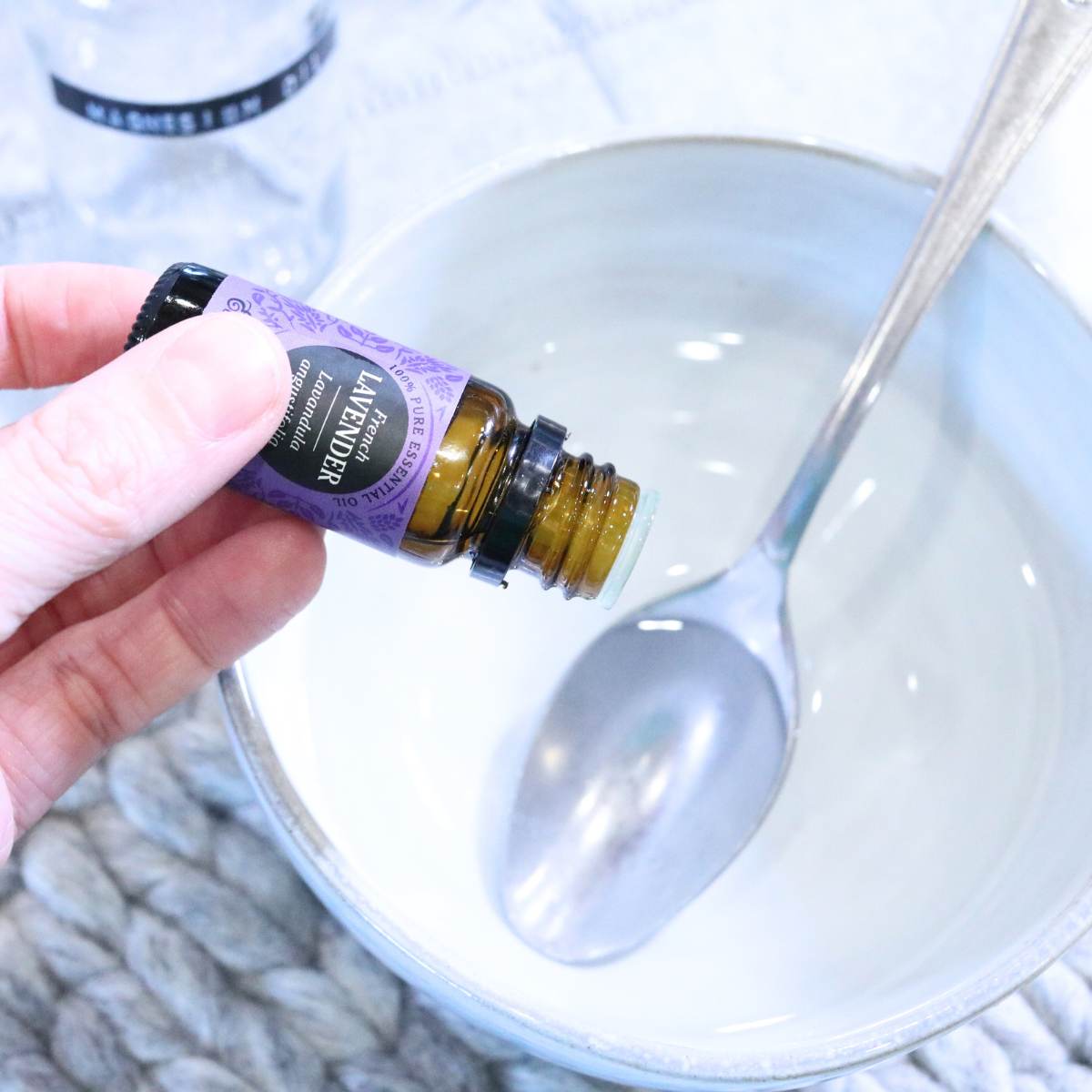 Carefully adding fragrance to her homemade magnesium oil, a woman is adding drops of French Lavender the bowl of other ingredients. A silver spoon is resting in the bowl to stir when she is finished.