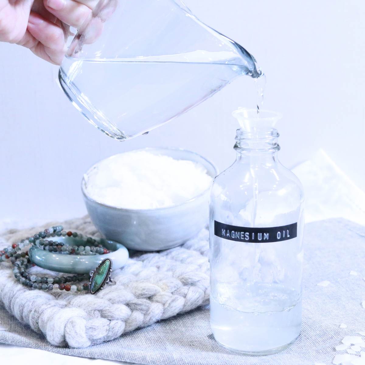 A hand is seen holding onto a clear glass pitcher with a pour spout carefully pouring a magnesium oil into a glass storage bottle. A stack of turquoise bracelets and a jade bracelet can be seen in the background of the photo. Magnesium flakes are scattered around the tabletop.