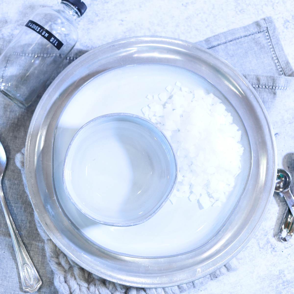 A table containing supplies for DIY magnesium oil recipe. A plate with white flaked magnesium flakes, a small bowl filled with distilled water, silver measuring spoons and a clear, empty bottle labeled magnesium oil ready to make the recipe.