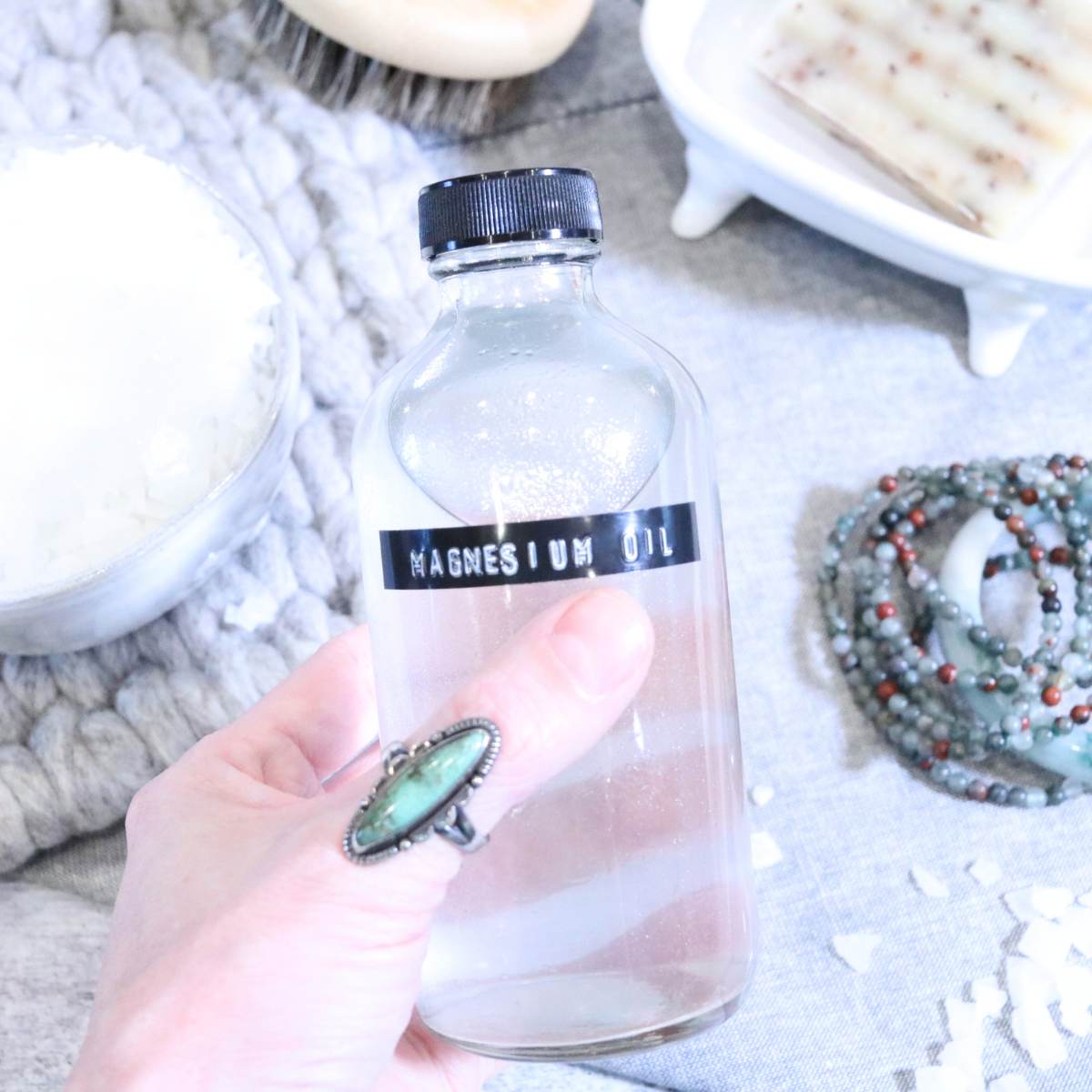 A womans hand wearing a turquoise ring is seen holding onto a clear glass bottle of homemade magnesium oil. In the background of the photo you can see a small stack of turquoise bracelets, a bowl of magnesium flakes used in the recipe, a dry brush and a bar of homemade patchouli soap. Magnesium flakes are scattered about around the table top.