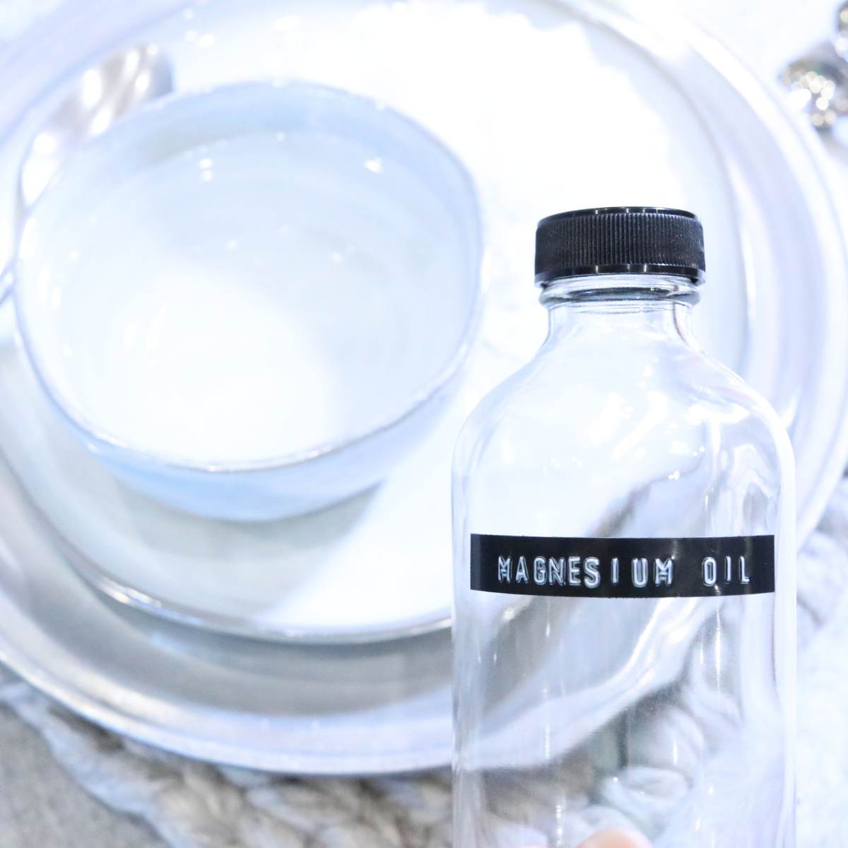 An example of how to store magnesium oil. 8 ounce clear jar with black lid and embossed label are sitting empty along with a bowl of boiling water and a plate of magnesium flakes. 