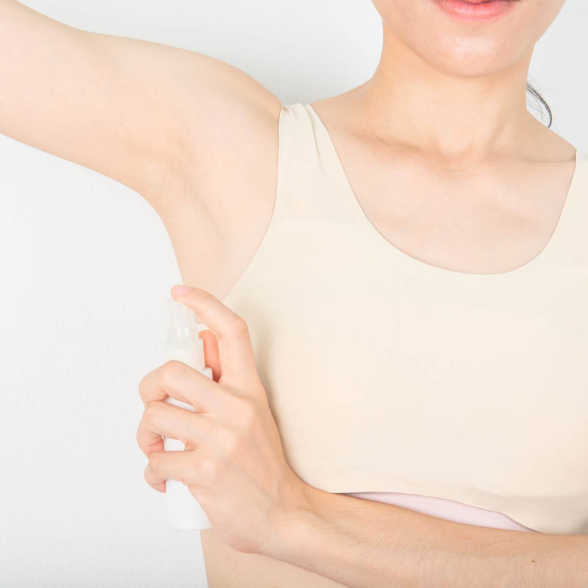 A woman wearing a cream colored sports top is demonstrating how to use magnesium deodorant spray. The deodorant spray bottle she is holding is small and white with a clear spray top.