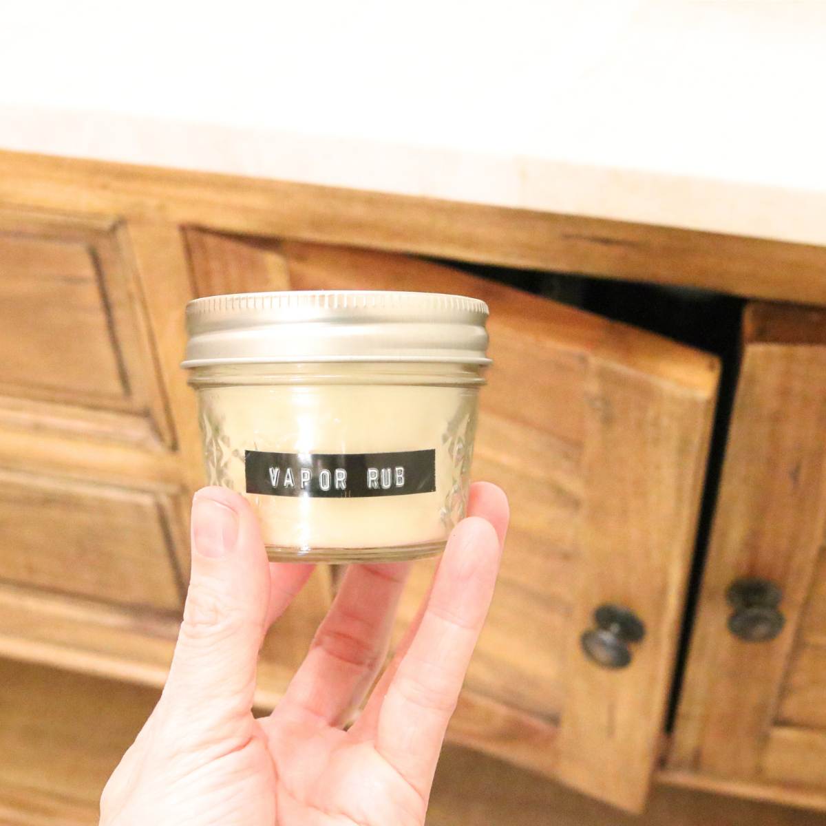 A woman is holding a clear glass jar of homemade vapor rub that is neatly labeled with an embossing label maker. She is holding up the jar in front of her wooden bathroom cabinet to show how the vapor rub should be stored if it is in a clear glass jar instead of an amber colored jar. Essential oils should be kept in a cool dark place away from sunlight. The vapor rub recipe contains essential oils.