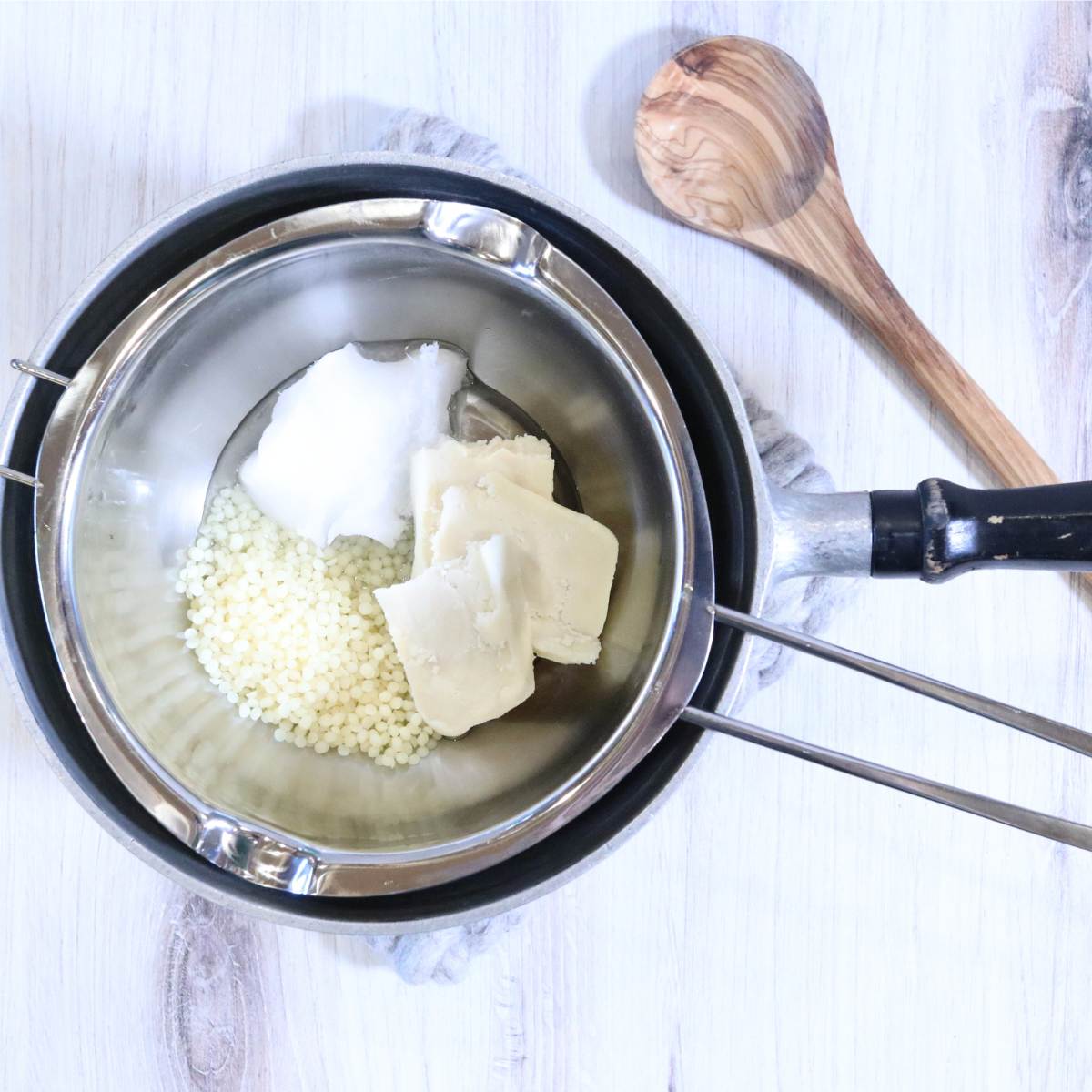 A double boiler pot sits with ingredients in it for an all natural vapor rub recipe. A wooden spoon sits nearby to stir when the ingredients begin to melt.