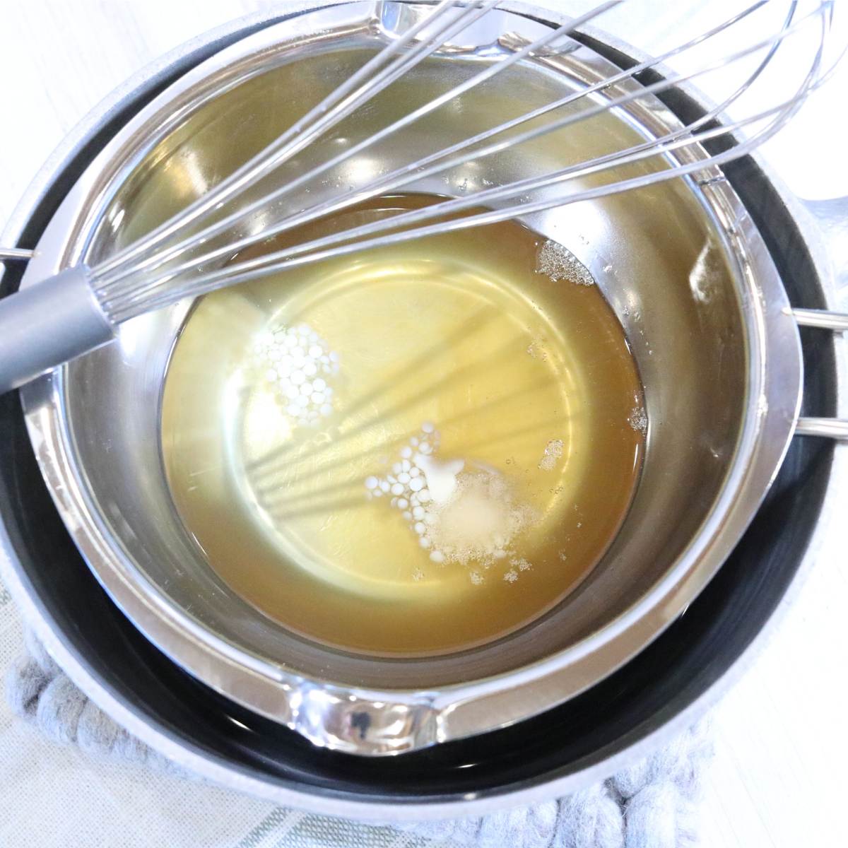 A clear yellow liquid sitting in a double boiler pot. A trace amount of beeswax pellets can be seen in the liquid that have not yet melted. A silver whisk sits ontop of the pot ready to whisk ingredients together.