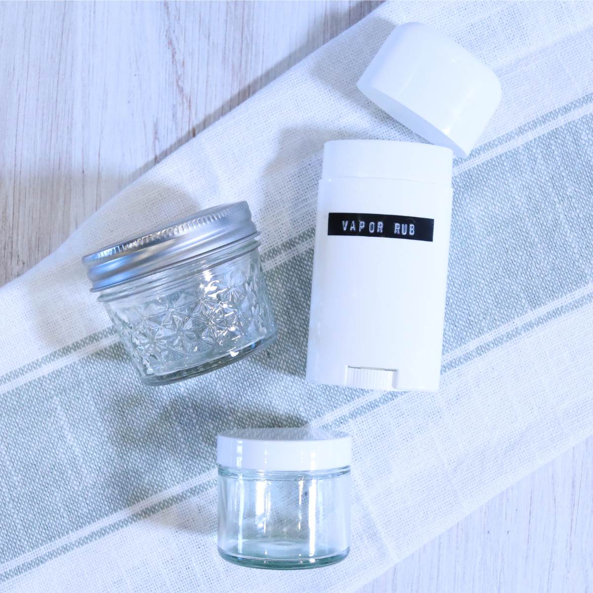 Three separate containers that can be used to store homemade all-natural vapor rub. A white empty deodorant container labeled Vapor Rub, and two smaller clear glass jars with airtight lids are resting on a light green and white striped hand towel.