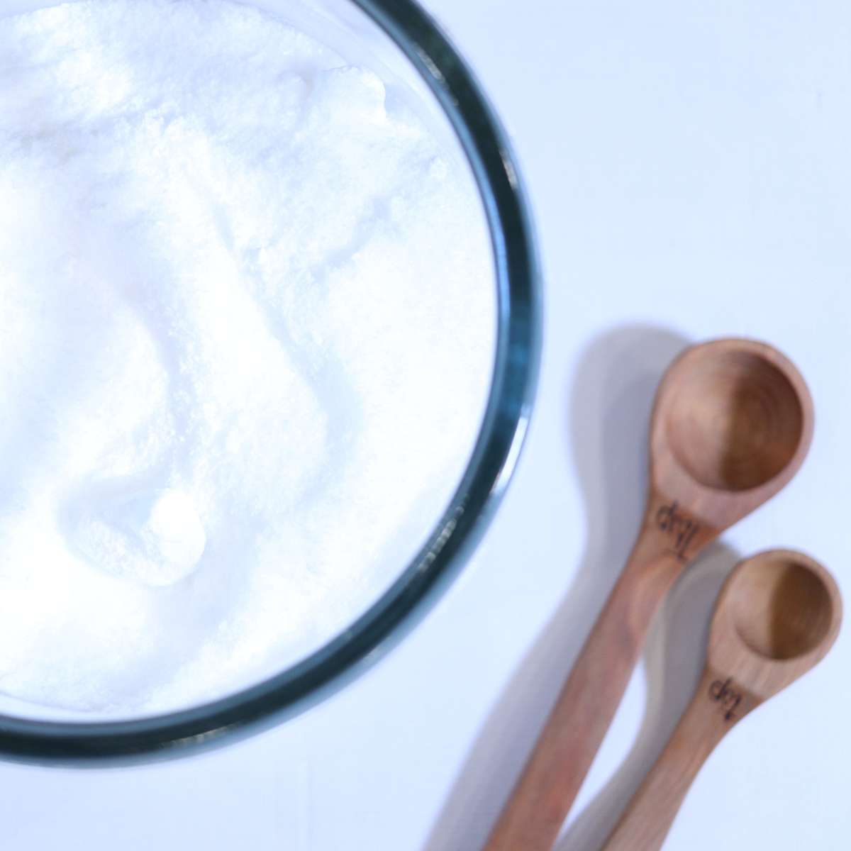 White powdered laundry scent booster in a clear glass jar with two wooden measuring spoons nearby.