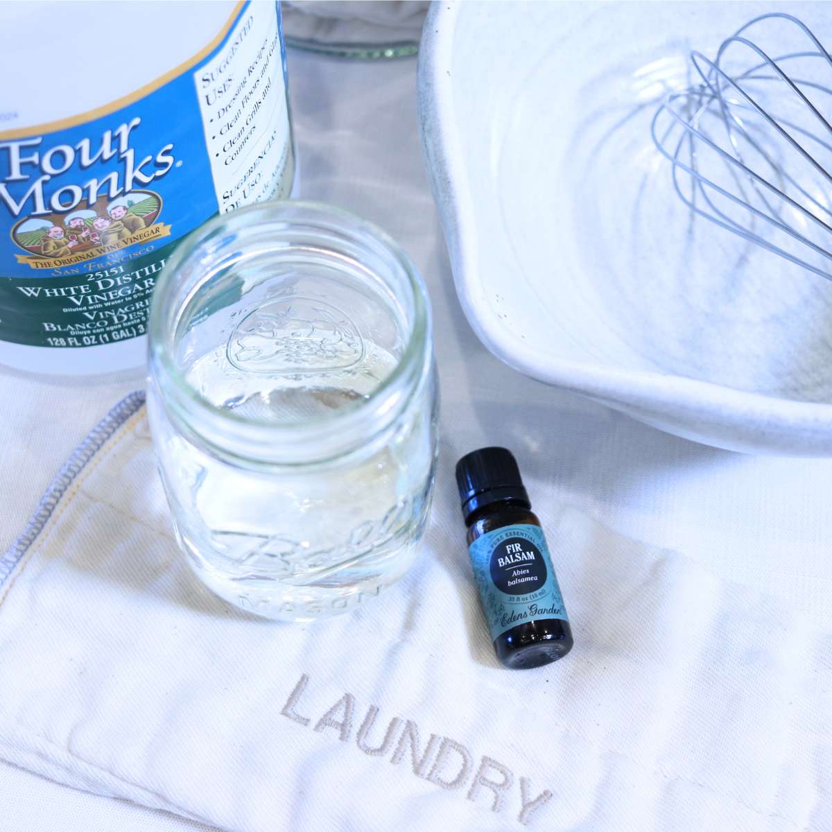 Ingredients that will be used to make homemade non-toxic dryer sheets. A bottle of white vinegar, filtered water in a clear glass, a mixing bowl and metal whisk and the cloth pieces for the dryer sheets are resting on a table. An amber bottle of fir balsam essential oil will be used to add fragrance to the homemade dryer sheets.