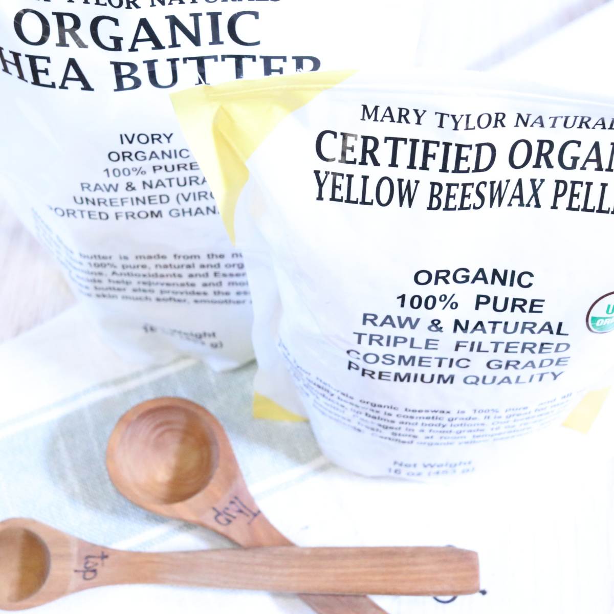 Two bags of ingredients being used for a DIY vapor rub recipe are resting on a tabletop with wooden measuring spoons sitting in front. A yellow and white bag of shea butter and beeswax pellets can be seen in the photo.