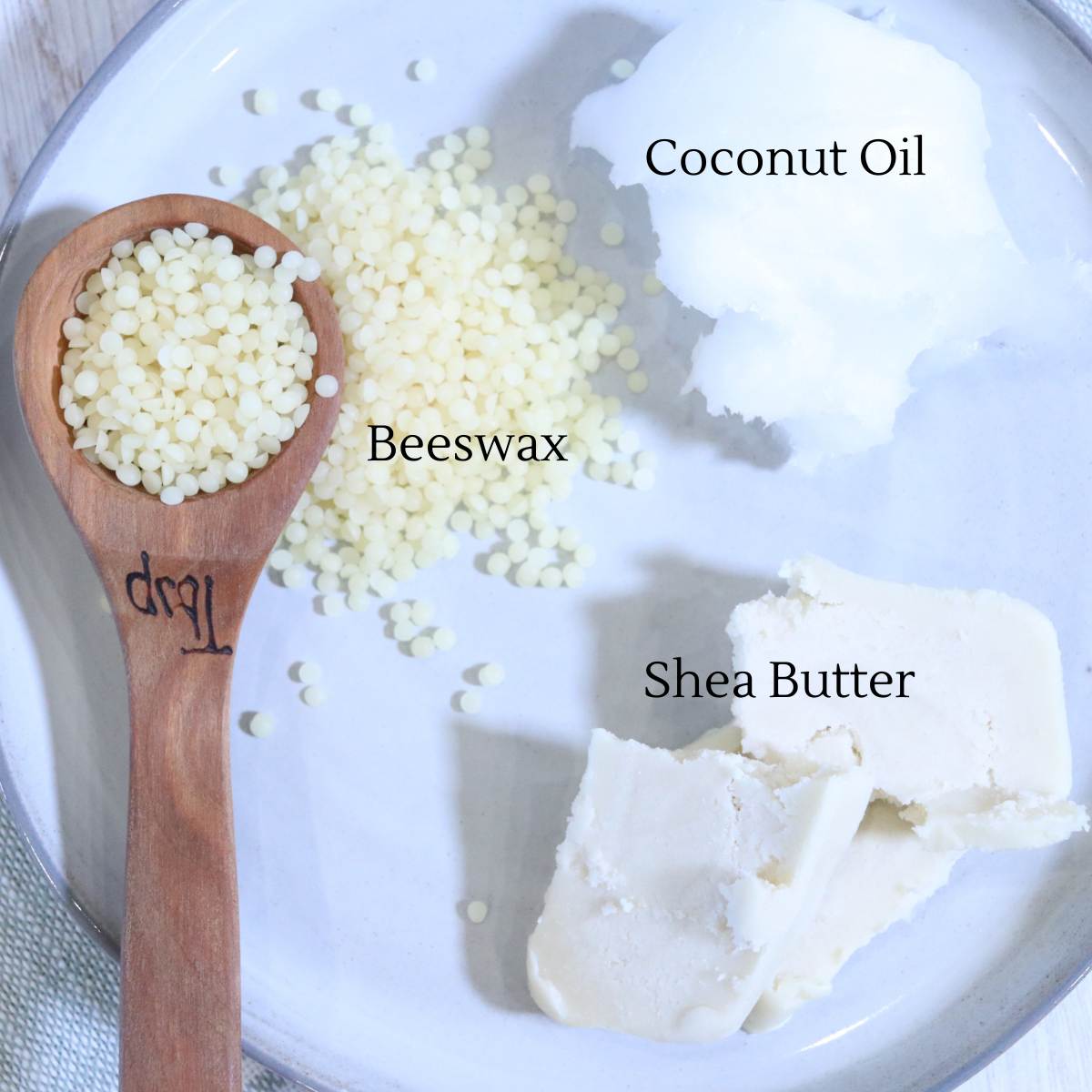 A small plate of ingredients for homemade vapor rub recipe. The plate has shea butter, beeswax pellets and coconut oil on it. A small wooden measuring spoon also rests on the plate.