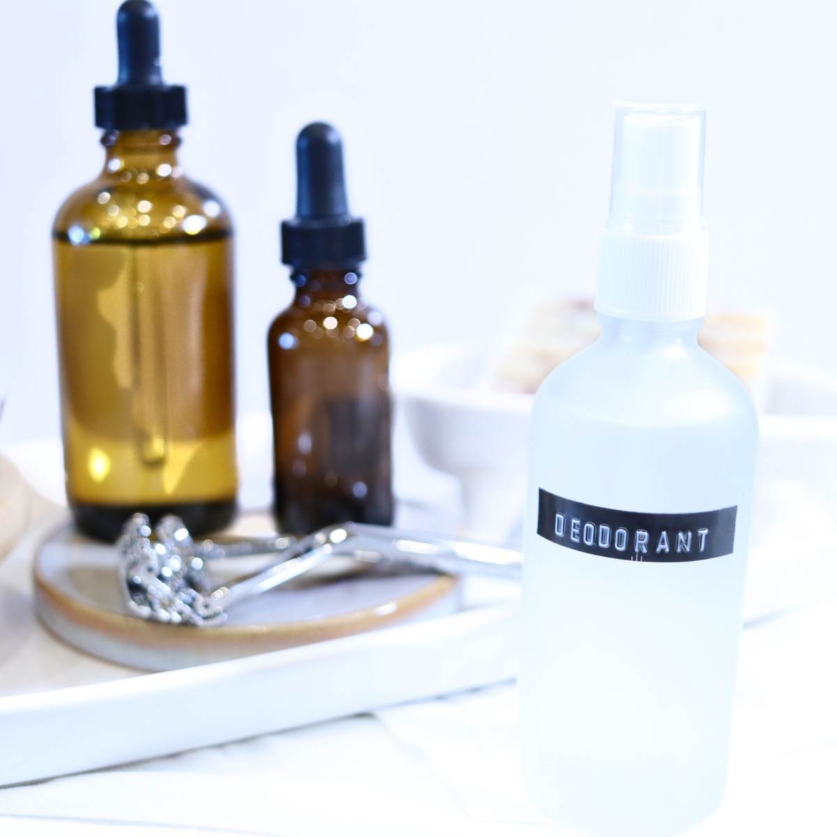 A picture of homemade magnesium deodorant spray in a clear glass bottle with white spray cap. Two larger amber glass dropper bottles full of other oils is in the background along with a silver zero waste razor.