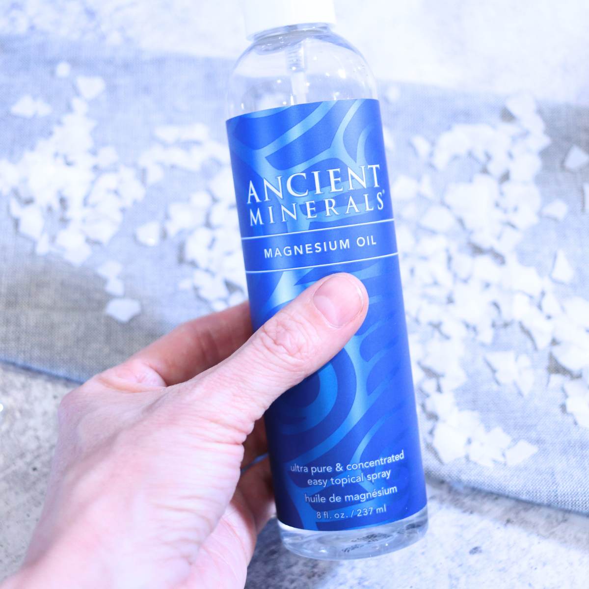 A Womans hand is holding a clear plastic bottle with a bright multicolored blue wrapper filled with magnesium oil. The oil will be used with other ingredients to make DIY magnesium deodorant. In the background of the picture, magnesium flakes are scattered about onto of a gray tablecloth.