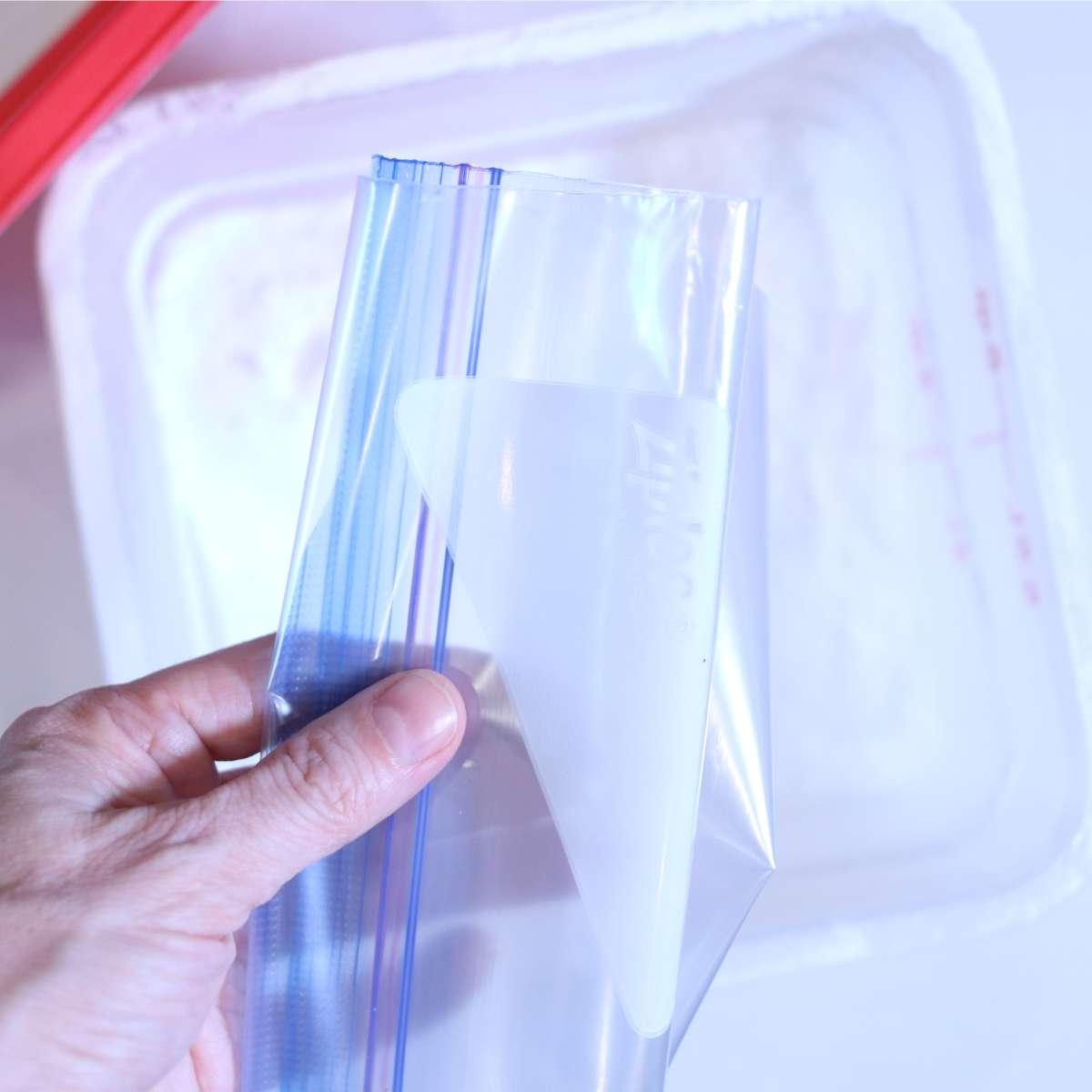 A woman hand is holding a folded clear ziplock bag in front of a large container filler with a homemade laundry scent booster powder. She is sharing multiple ways to combine ingredients without the mess. 