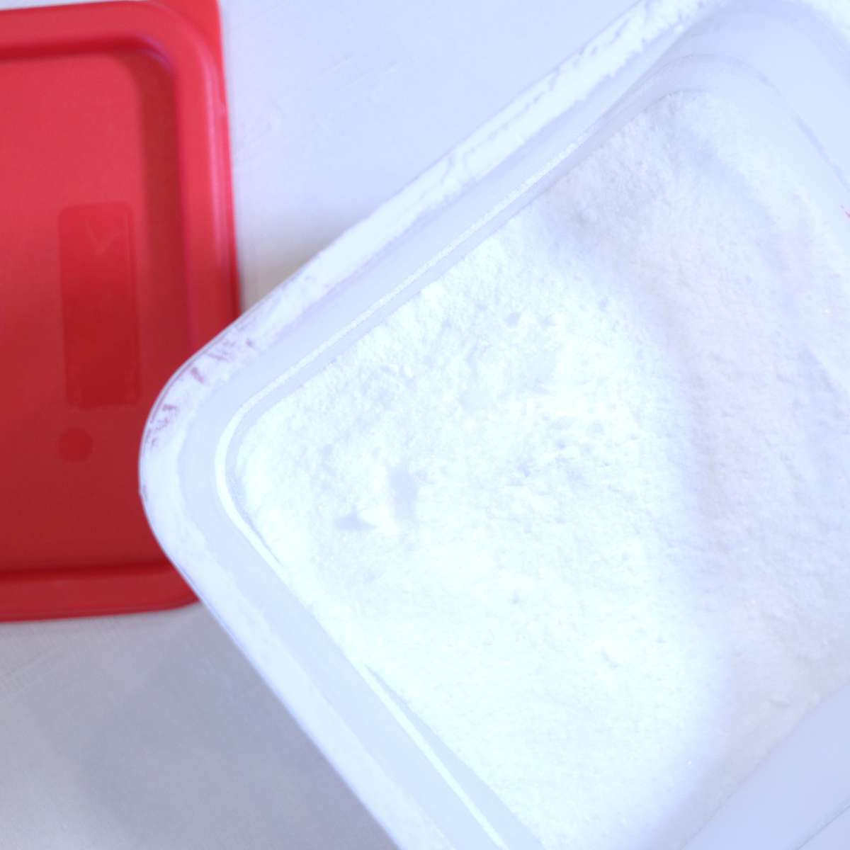 A large plastic container with a red lid are sitting on the table filled with ingredients to make a large batch of homemade scent booster. 