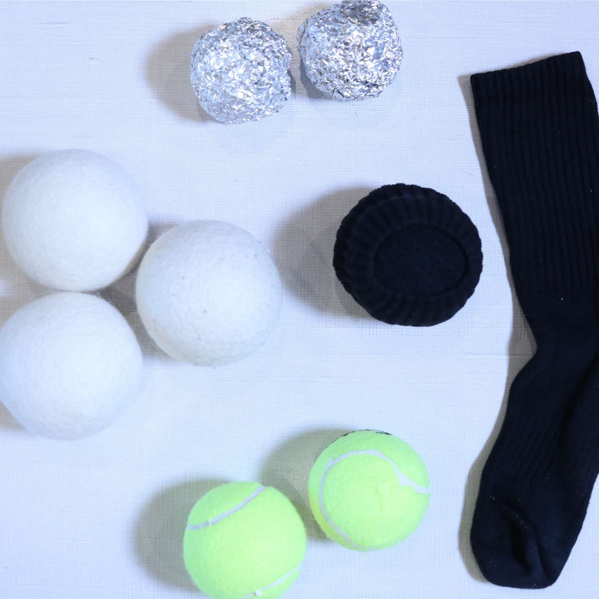 A table top with multiple DIY dryer ball laundry hacks including tennis balls, old socks shaped into a ball and 2 large balls of aluminum foil.