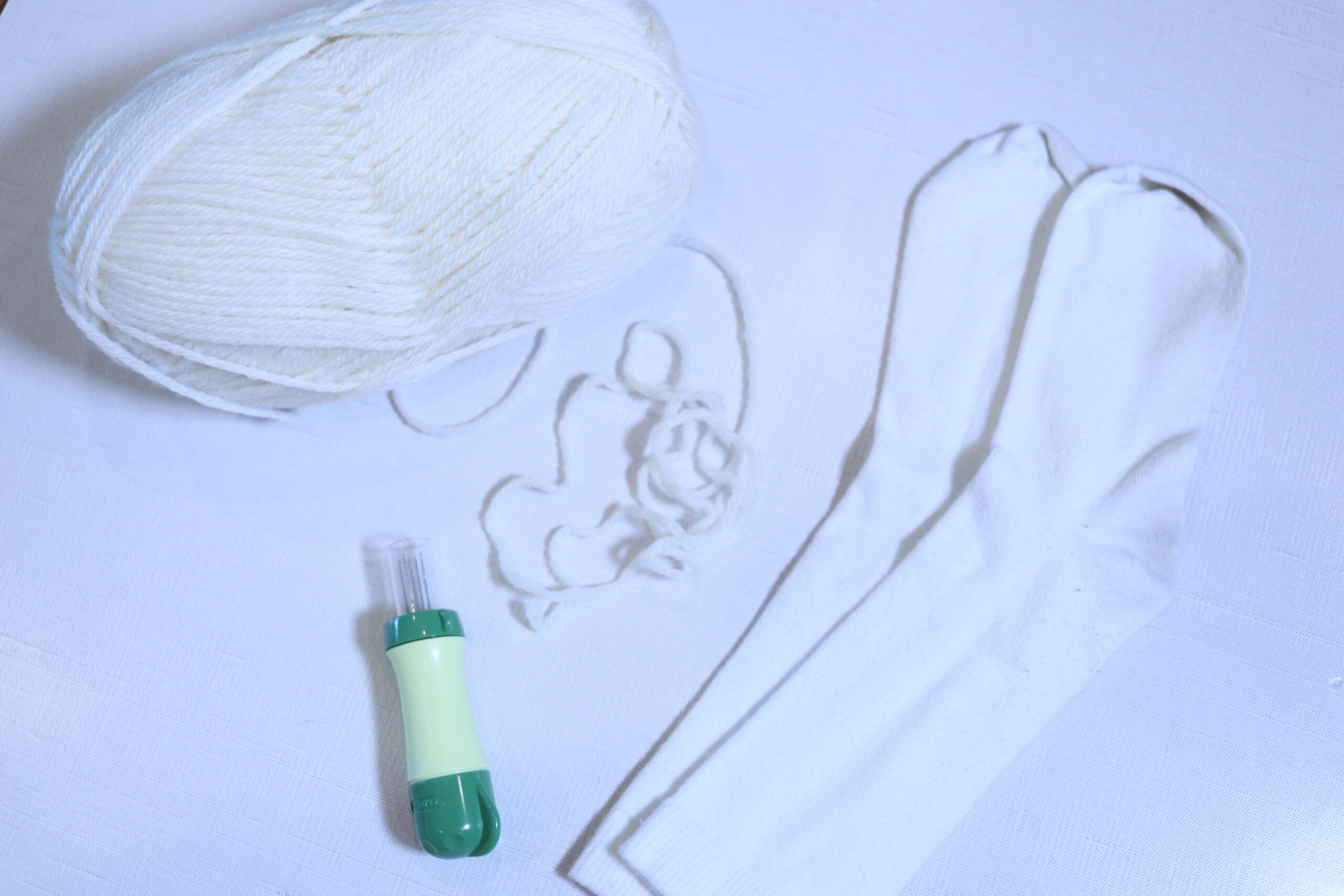 A white table top with a skein of cream wool yarn, a wool felting tool and a pair of old white socks that will be used for making DIY wool dryer balls.