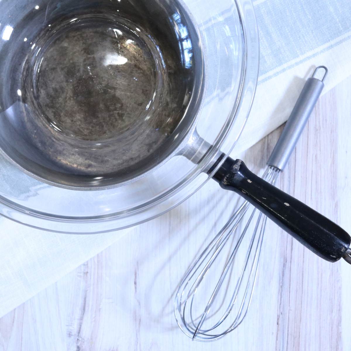 A homemade double boiler can be easily made using a pot with a glass bowl resting on top. A metal whisk can be seen sitting next to the double boiler pot being used for a homemade vapor rub recipe.