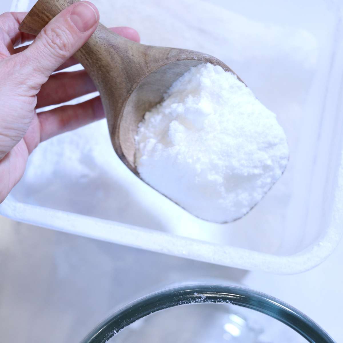 A womans hand is holding a wooden scoop that contains white powdered scent booster. She is transferring the mixture to a clear glass storage jar so it is ready to be used.