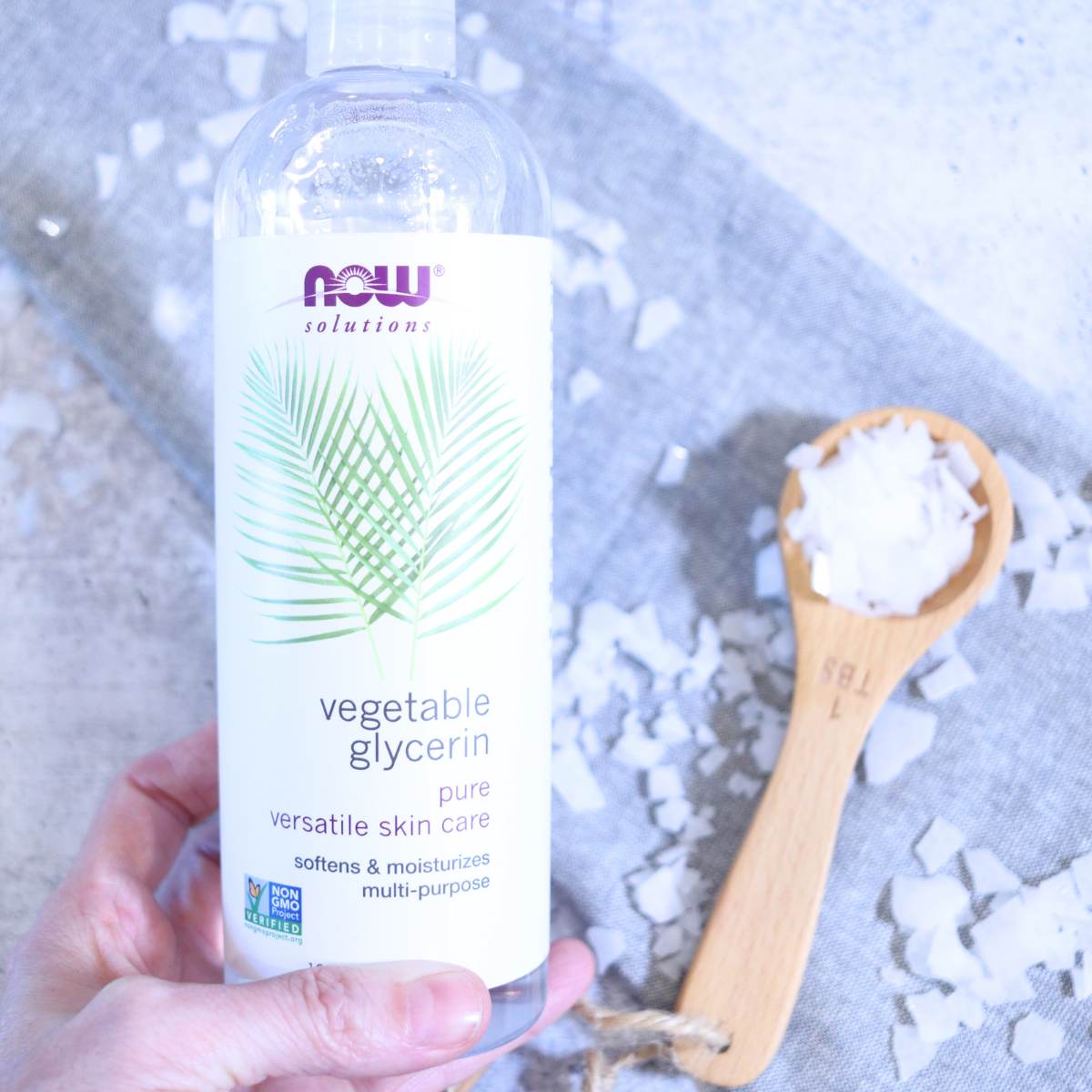 Holding onto a clear plastic bottle filled with clear vegetable glycerin, a woman is preparing to use for her homemade magnesium deodorant recipe. A wooden Tablespoon filled with magnesium flakes is sitting on the side while other magnesium flakes are seen scattered throughout the photograph.