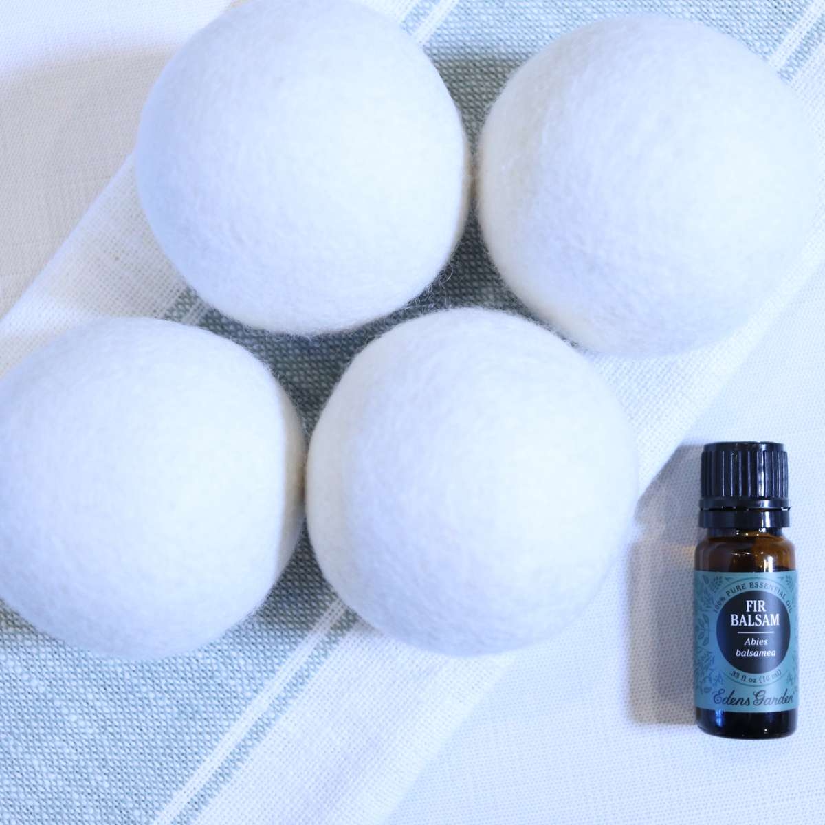 Four white wool felted dryer balls resting on a kitchen towel folded neatly on the table. A bottle of fir essential oil sitting nearby to add fragrance to the dryer balls.