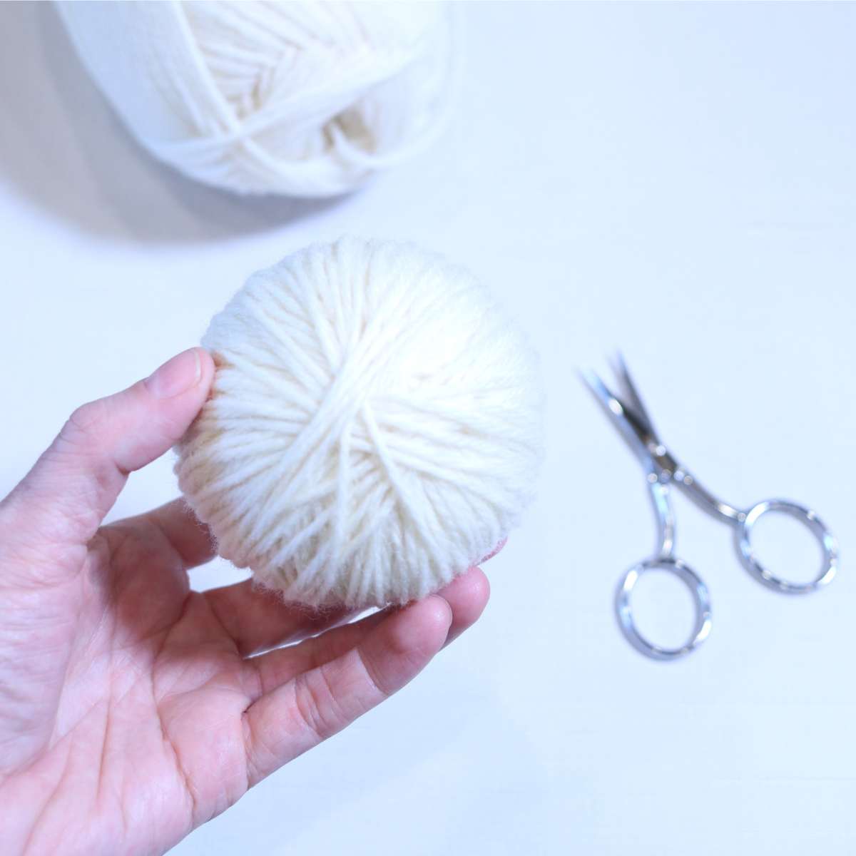 A ball of cream yarn and silver scissors rests on a table while a womans hand is holding up a completed wool dryer ball she just finished making.